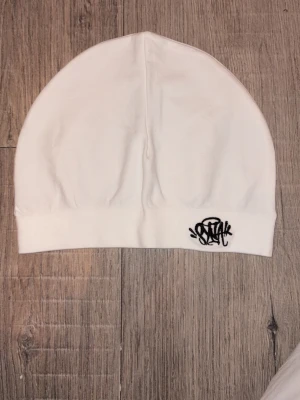 Vit syna world skull cap med svart broderad logga - Snygg vit syna world skull cap med svart broderad SNAK-logga framtill. Enkel och stilren design i mjukt tyg som sitter skönt på huvudet. Perfekt accessoar för att lyfta din streetwear-look.