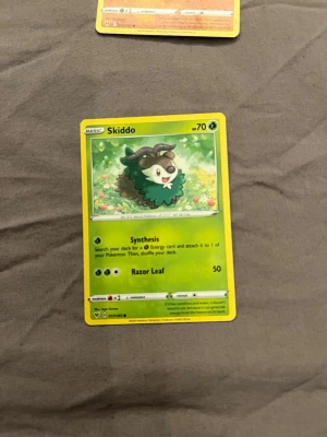 Pokémonkort Skiddo grön/gul - Säljer ett Pokémonkort med Skiddo-motivet. Kortet har en grön och gul ram, med Skiddo illustrerad på en blomsteräng. Text och symboler är tryckta i svart och vitt. Perfekt för samlare eller Pokémonfans.