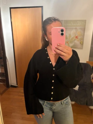 Svart kofta från Gina Tricot - Säljer en svart kofta från Gina Tricot med v-ringning och dekorativa knappar framtill. Koftan har vida ärmar och ribbad nederkant som ger en snygg siluett. Perfekt att matcha med jeans eller kjol för en trendig look.💕