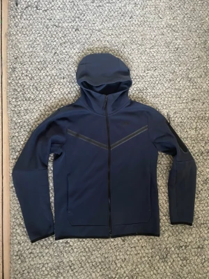 Mörkblå Nike Tech Fleece hoodie - Snygg mörkblå Nike Tech Fleece hoodie med hel dragkedja och huva. Hoodien har svarta detaljer över bröstet och på ärmen, samt två fickor framtill. Tillverkad i ett mjukt och stretchigt material som passar perfekt till en sportig och avslappnad stil.