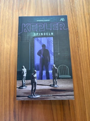 Kepler - Spindeln kriminalroman - Spännande kriminalroman 'Spindeln' av Kepler. Boken har ett mörkt omslag med lila text och figurer i grått, och är utgiven av Albert Bonniers Förlag. Perfekt för dig som gillar nervkittlande deckare och Joona Linna-serien.
