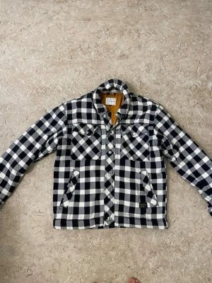 Svartvit rutig overshirt från Carhartt - Overshirt i svartvitt rutigt mönster från Carhartt. Jackan har två bröstfickor med lock, snedställda sidfickor och knäppning framtill. Fodrad med kontrasterande gul insida. Perfekt för lager-på-lager och streetwear-stil.