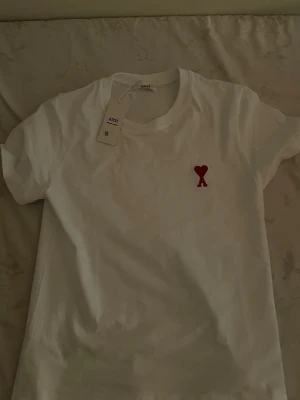 Vit t-shirt från Ami med rött hjärta - Vit t-shirt från Ami Alexandre Mattiussi med ett litet rött hjärta broderat på bröstet. Klassisk rund halsringning och korta ärmar. Perfekt basic-plagg med en snygg detalj framtill.