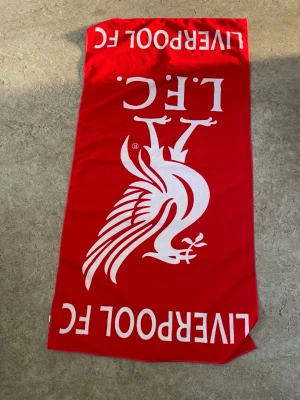 Röd Liverpool FC halsduk - Röd halsduk med stort vitt Liverpool FC-tryck och Liverbird-logga. Perfekt accessoar för alla fans som vill visa sitt stöd för laget. Halsduken har en klassisk rektangulär form och är tillverkad i mjukt material.