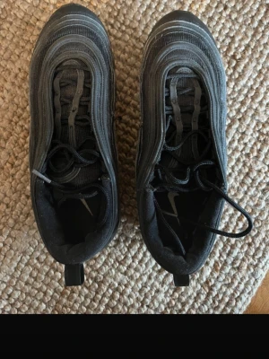Nike Air Max 97 svarta sneakers - Nike Air Max 97 sneakers i helsvart färg med klassisk vågig design och synlig Air-sula. Skorna har snörning, rund tå och platt sula. Materialet är en mix av syntet och mesh för skön komfort. Snyggt Nike-märke på plösen och innersulan. Perfekt för dig som gillar streetstyle.
