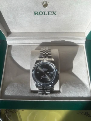 Rolex Datejust silver klocka - Snygg Rolex Datejust med silverfärgad länk i rostfritt stål och svart urtavla. Klockan har räfflad bezel och klassisk jubilee-länk. Levereras i originalbox med certifikat och tillbehör. Perfekt för dig som vill ha en ikonisk och tidlös accessoar.