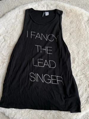 Svart linnetopp från Divided H&M - Svart ärmlös linnetopp från Divided by H&M med vit text 'I FANCY THE LEAD SINGER' framtill. Toppen har en loose passform, perfekt för en avslappnad och cool stil. Obs mindre i strl skulle säga passar S 