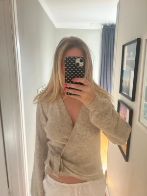 Beige omlottkofta i ribbstickat - Snygg beige omlottkofta i ribbstickat material med knytband i sidan. Koftan har långa ärmar och en djup v-ringning, perfekt för lager på lager. Enkel och stilren design som passar till det mesta.