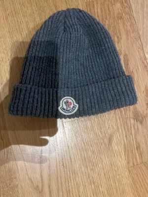 Moncler mössa - Säljer en grå ribbstickad mössa från Moncler med uppvikt kant och klassisk Moncler-logga framtill. Mössan är tillverkad i mjukt material och har en enkel, stilren design som passar perfekt till höstens och vinterns outfits.