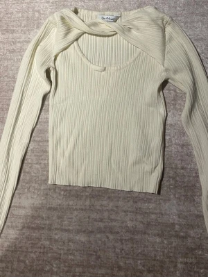 Ribbad cremefärgad topp från NA-KD - Trendig ribbad topp i cremevitt från NA-KD med lång ärm och unik twistad detalj vid halsen. Toppen har en croppad passform och rundad halsringning. Perfekt för dig som gillar stilrena men ändå speciella plagg. Kan ändra pris om ni kontaktar mig 