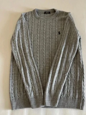 Grå kabelstickad tröja Polo Ralph Lauren - Kabelstickad grå tröja från Polo Ralph Lauren med rund halsringning och svart broderad logga på bröstet. Tröjan har långa ärmar och ribbade muddar vid ärmslut och nederkant. Perfekt för lager-på-lager och klassisk stil.