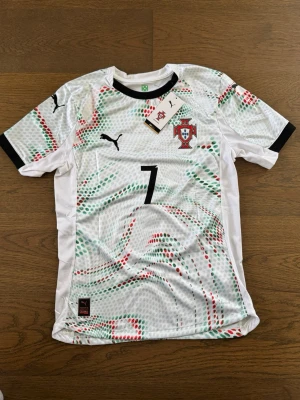Portugal matchtröja Puma Ronaldo #7 - Snygg vit Portugal matchtröja från Puma med svarta detaljer, nummer 7 och Ronaldo på ryggen. Tröjan har gröna och röda grafiska mönster, rund hals och korta ärmar. Perfekt för dig som vill stötta landslaget eller bära en ikonisk fotbollströja.