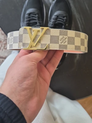 Vit och grå LV-bälte med rutigt mönster - Snyggt bälte från Louis Vuitton i vitt och grått med klassiskt rutigt mönster och stor LV-logga i guld som spänne. Perfekt accessoar för att lyfta din outfit med en exklusiv touch. Tillverkat i skinn med tydlig Louis Vuitton Paris-text på bandet.