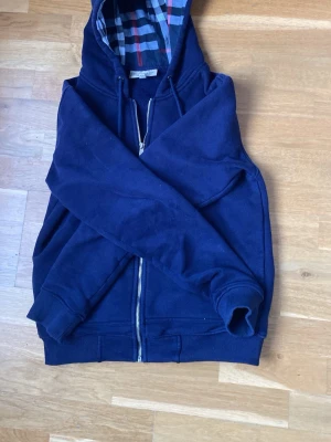 Mörkblå hoodie med dragkedja helt ny knappt använd  - Mörkblå hoodie med dragkedja och huva med rutigt innerfoder. Två fickor framtill och ribbade muddar vid ärmslut och nederkant. Snygg och enkel design, perfekt till jeans eller joggers.
