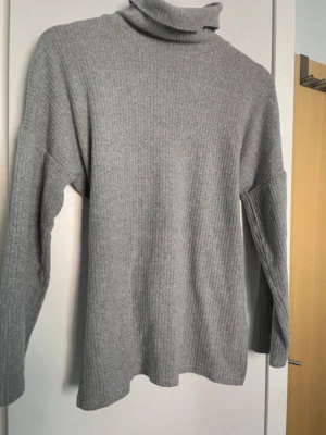 Grå ribbad tröja med hög hals & knytband - Mysig ribbad tröja i grå färg med hög hals. Har ett avtagbart/justerbart knytband i midjan som ger en fin siluett om man vill markera midjan. Funkar lika bra löst för en mer avslappnad stil.