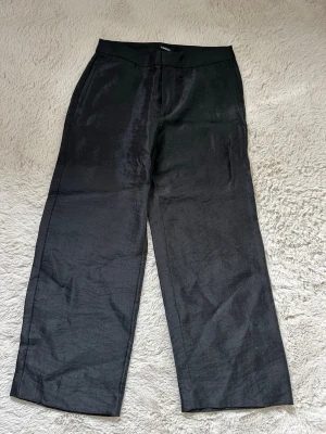 Svarta kostymbyxor från Lindex - Snygga svarta kostymbyxor från Lindex med rak passform och diskret glansigt tyg. Byxorna har hög midja, dold dragkedja och hyska fram. Perfekta för en stilren look och lätta att matcha med olika toppar.