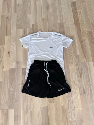 Nike träningsset t-shirt och shorts - Snyggt träningsset från Nike med en vit t-shirt i funktionsmaterial och svarta shorts med dragsko och reflexdetaljer. Båda plaggen har Nike-logga och är perfekta för gym eller löpning. T-shirten har rund hals och kort ärm, shortsen har elastisk midja.