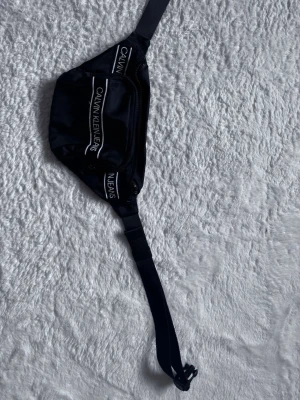 Svart magväska från Calvin Klein Jeans - Snygg svart magväska från Calvin Klein Jeans med justerbart band och dragkedja. Väskan har vita logotexter på framsidan och är tillverkad i ett slitstarkt syntetmaterial. Perfekt för att bära det viktigaste när du är på språng.