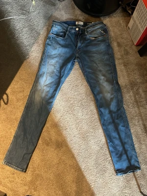 Blå slim fit jeans Replay anbass  - Säljer ett par blå slim fit jeans från Replay med klassisk femficksdesign och snygga slitningar. Jeansen har normal midja och smala ben, tillverkade i mjukt denimtyg för skön passform. Perfekta för en avslappnad och trendig look.