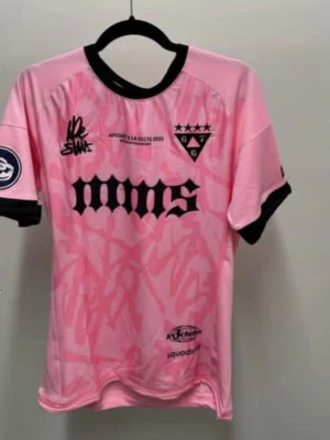 Rosa fotbollströja med svarta detaljer - Snygg rosa fotbollströja med svarta detaljer och tryck. Tröjan har svart krage, svarta muddar på ärmarna och flera logotyper på bröstet och ärmen. Tillverkad i ett lätt och ventilerande material, perfekt för träning eller match.
