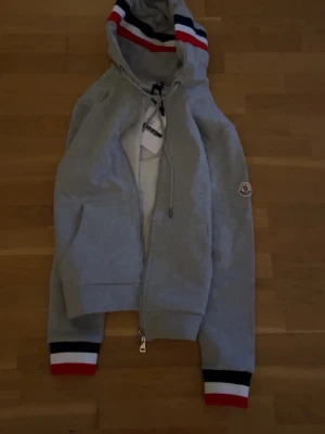 Grå Moncler hoodie med dragkedja - Snygg grå hoodie från Moncler med dragkedja framtill och huva med röda, vita och marinblå ränder. Ärmsluten har matchande ränder och Moncler-loggan sitter på ena ärmen. Perfekt för en sportig och trendig look.