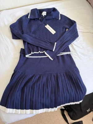 Marinblå stickad klänning från American 1985 - Marinblå stickad klänning med kabelstickat mönster, vit krage och vita detaljer vid midja och nederkant. Klänningen har långärmade ärmar, v-ringning och plisserad kjol. Perfekt för en preppy look.