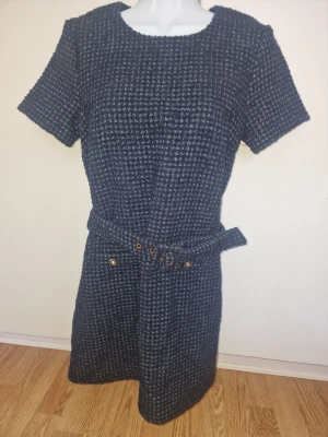 Mörkgrå tweedklänning med bälte - Snygg mörkgrå tweedklänning med kort ärm och rund halsringning. Klänningen har två fickor framtill med guldfärgade knappar och ett matchande bälte i midjan. Materialet är grovt vävt och ger en klassisk look.