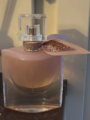 Lancome La Vie Est Belle EdP 30ml - Helt ny, endast sprayad för att känna doften 💕Elegant parfymflaska från Lancome i ljusrosa glas med glittrigt band och dekorativt blad runt halsen. Flaskan har en fyrkantig form och ett genomskinligt lock. Perfekt för dig som gillar lyxiga och feminina dofter.