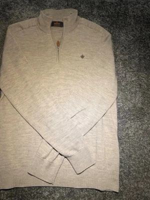 Beige halvzip tröja från Morris - Snygg beige stickad tröja från Morris med halvzip och diskret broderad logga på bröstet. Tröjan har ribbade muddar och krage, perfekt för lager-på-lager och en clean look.