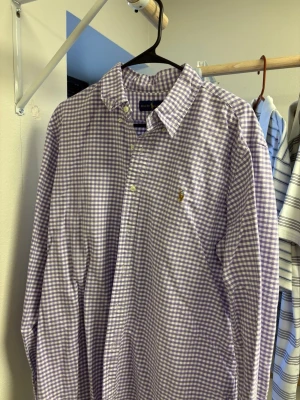 Lila/vit rutig skjorta från Ralph Lauren - Säljer en klassisk lila och vit rutig skjorta från Ralph Lauren med lång ärm och knappar framtill. Skjortan har en broderad logga på bröstet och är tillverkad i mjuk bomull. Perfekt för dig som gillar stilrena och tidlösa plagg.