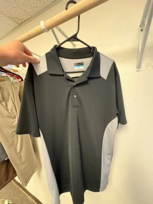 Svart och grå polotopp från PGA Tour - Snygg svart och grå polotopp från PGA Tour med klassisk krage och två knappar. Materialet är polyester och designen har kontrasterande grå paneler på axlar och sidor. Perfekt för dig som gillar sportig stil.