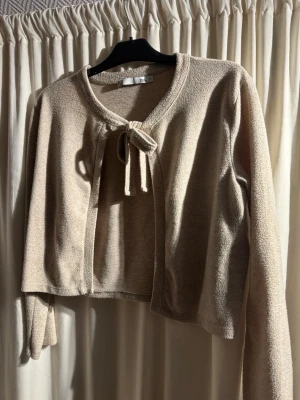 Beige kofta med knyt från 157 - Snygg beige kofta från 157 med knytband framtill. Koftan är croppad och har långa ärmar samt en mjuk, lite texturerad yta. Perfekt att slänga över en topp.