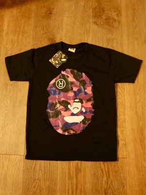Svart BAPE t-shirt med camo-tryck - Skickar snabbt📦 | Ny med tags👔| Cool svart t-shirt från BAPE med stort färgglatt camo-tryck i lila, rosa och blå toner på bröstet. Klassisk rund hals och korta ärmar. Perfekt för dig som gillar streetwear och vill sticka ut med en ikonisk design.