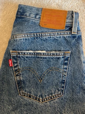Levi's 501 blå raka jeans - Klassiska blå Levi's 501 jeans med raka ben och mid-waist midja. Jeansen har den ikoniska röda Levi's-lappen på bakfickan och läderpatch i midjan. Tillverkade i slitstark denim med kontrastsömmar och femficksmodell. Perfekta för en avslappnad och tidlös stil.