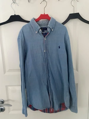 Ljusblå skjorta från Ralph Lauren - Klassisk ljusblå skjorta från Ralph Lauren med button-down krage och broderad logga på bröstet. Skjortan har långa ärmar och knappar framtill. Tillverkad i mjuk bomull som ger en avslappnad men ändå stilren look.