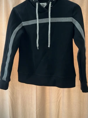 Svart hoodie från Maya Delorez - Snygg svart hoodie från Maya Delorez med gråa detaljer längs ärmar och bröst samt gråa dragsnören i huvan. Märkesnamnet står tryckt på huvkanten. Hoodien har en avslappnad passform och är tillverkad i mjukt material, perfekt för en chill look.
