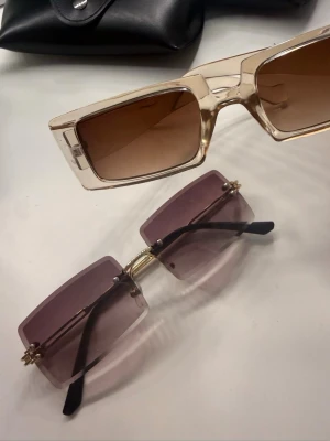 Två par trendiga solglasögon - Säljer två par solglasögon med rektangulära bågar. Ett par har transparent beige båge och bruna glas, det andra har guldiga detaljer och lila tonade glas utan båge runt. Perfekta accessoarer för en cool look.