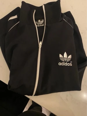 Svart Adidas hoodie med dragkedja - Säljer en svart hoodie från Adidas med vit dragkedja och vita detaljer. Klassisk Adidas-logga broderad på bröstet och ribbade muddar vid ärmsluten. Hoodien har hög krage och är tillverkad i ett mjukt, sportigt material. Perfekt för en avslappnad och sportig stil.