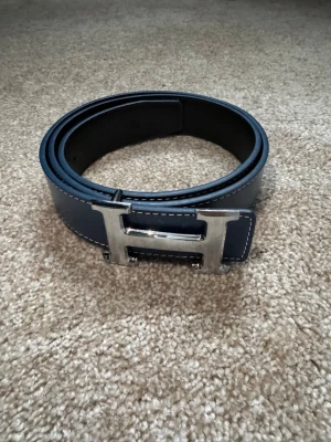 Blått skärp med H-spänne - Snyggt blått skärp med ett stort, silverfärgat H-spänne. Skärpet har vita sömmar längs kanterna och är gjort i skinn. Perfekt accessoar för att lyfta din outfit. Helt oanvänt.