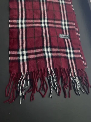 Rutig vinröd halsduk från Burberrys - Klassisk halsduk i vinrött med rutigt mönster i svart, vitt och rosa från Burberrys. Tillverkad i 100% lammull och har fransar i ändarna. Perfekt accessoar för att lyfta din outfit med en tidlös brittisk vibe. Inga märkbara defekter. Pris går att diskutera!