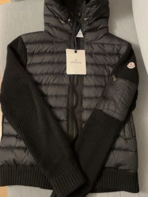 Svart Moncler cardigan med huva - Svart cardigan från Moncler med stickade ärmar och huva. Jackan har dragkedja framtill, ribbade muddar och en ficka med dragkedja på ärmen. Märkespatch på armen och quiltad front. Perfekt för dig som vill ha en snygg och varm jacka med street vibe. Köp och få ett par nuddie jeans på köpet finns i min profil.