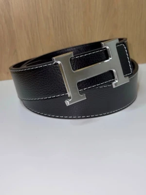 H-Bälte - Snyggt bälte med ett stort, silverfärgat H-spänne. Bältet är 115cm, har vit kontrastsöm längs kanterna och en klassisk, stilren design som lyfter vilken outfit som helst. Bältet är oanvänt (nytt). Du får även med ett armband och ett verktyg för att göra egna hål! Skriv vid minsta lilla fundering!