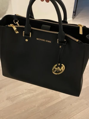 Svart handväska från Michael Kors - Snygg svart handväska från Michael Kors i slätt läder med guldfärgade detaljer och logotyp. Väskan har dubbla handtag, flera fack med dragkedja och en dekorativ MK-berlock framtill. Perfekt för dig som gillar stilrena accessoarer.
