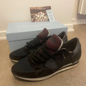 Philippe Model svarta sneakers - Snygga svarta sneakers från Philippe Model med detaljer i mocka, skinn och textil. Skorna har vinröd plös, svarta snören och en vit rand på sulan. Klassisk rund tå och quiltad häl för extra stilpoäng. Perfekta för dig som gillar premium och streetstyle. Endast skorna medföljer