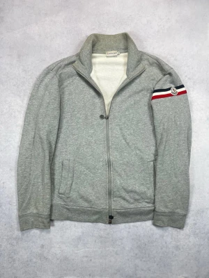 Grå zip-hoodie från Moncler - Snygg grå hoodie från Moncler med dragkedja framtill och två fickor. Ärmen har en röd, vit och blå rand med Moncler-logga. Mjuk bomullskänsla och hög krage för extra stilpoäng.