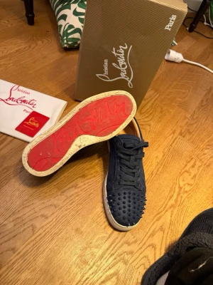 Blå Christian Louboutins - Snygga blå sneakers från Christian Louboutin med klassisk röd sula och coola nitar framtill. Skorna har snörning och är tillverkade i mocka för en lyxig känsla. Perfekta för dig som vill sticka ut med din stil. Har tappat en del spikar skick 6/10