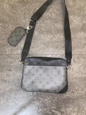 Louis Vuitton axelväska monogram grå ( pris kan diskuteras ) - Snygg axelväska från Louis Vuitton i grått och svart med klassiskt monogrammönster. Väskan har justerbar axelrem med logga och en liten avtagbar miniväska. Tillverkad i tåligt lädermaterial och har dragkedja upptill. Perfekt för dig som gillar streetstyle och lyx.