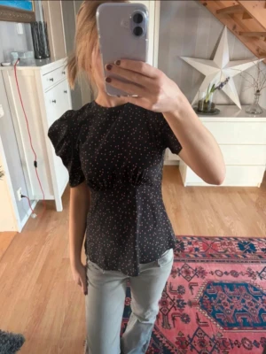Svart blus med puffärm och prickar - Supersöt svart blus med rosa prickar. Blusen har puffiga korta ärmar och markerad midja som ger en feminin siluett. Perfekt till jeans eller kjol för en trendig look. Materialet känns mjukt och följsamt.
