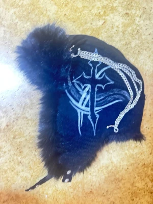 Svart trappermössa med kors och kedjor - Cool svart trappermössa med fuskpäls, broderat vitt korsmotiv och silvriga kedjor på sidan. Mössan har öronlappar och knäppning under hakan. Perfekt för dig som gillar alternativ stil och vill sticka ut.