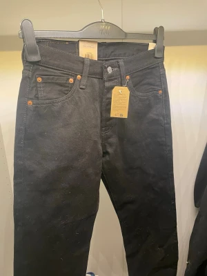 Svarta Levi's 501 jeans - Klassiska svarta Levi's 501 jeans med raka ben och knappgylf. Jeansen har fem fickor, nitar i kopparfärg och den ikoniska läderlappen bak i midjan. Tillverkade i slitstark denim med en tidlös look som alltid funkar.